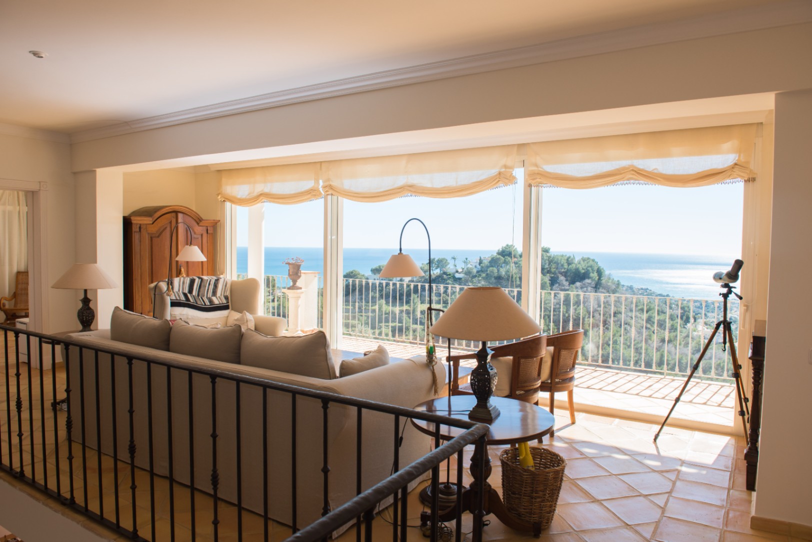 Lichte woonkamer met terracotta vloeren, smeedijzeren balustrade en grote ramen met panoramisch uitzicht op de Middellandse Zee en de kust.