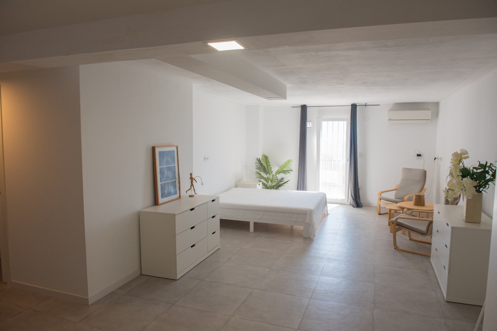 Minimalistische slaapkamer in neutrale tinten met grijze keramische vloer, functioneel wit meubilair, airconditioning en directe toegang naar buiten.