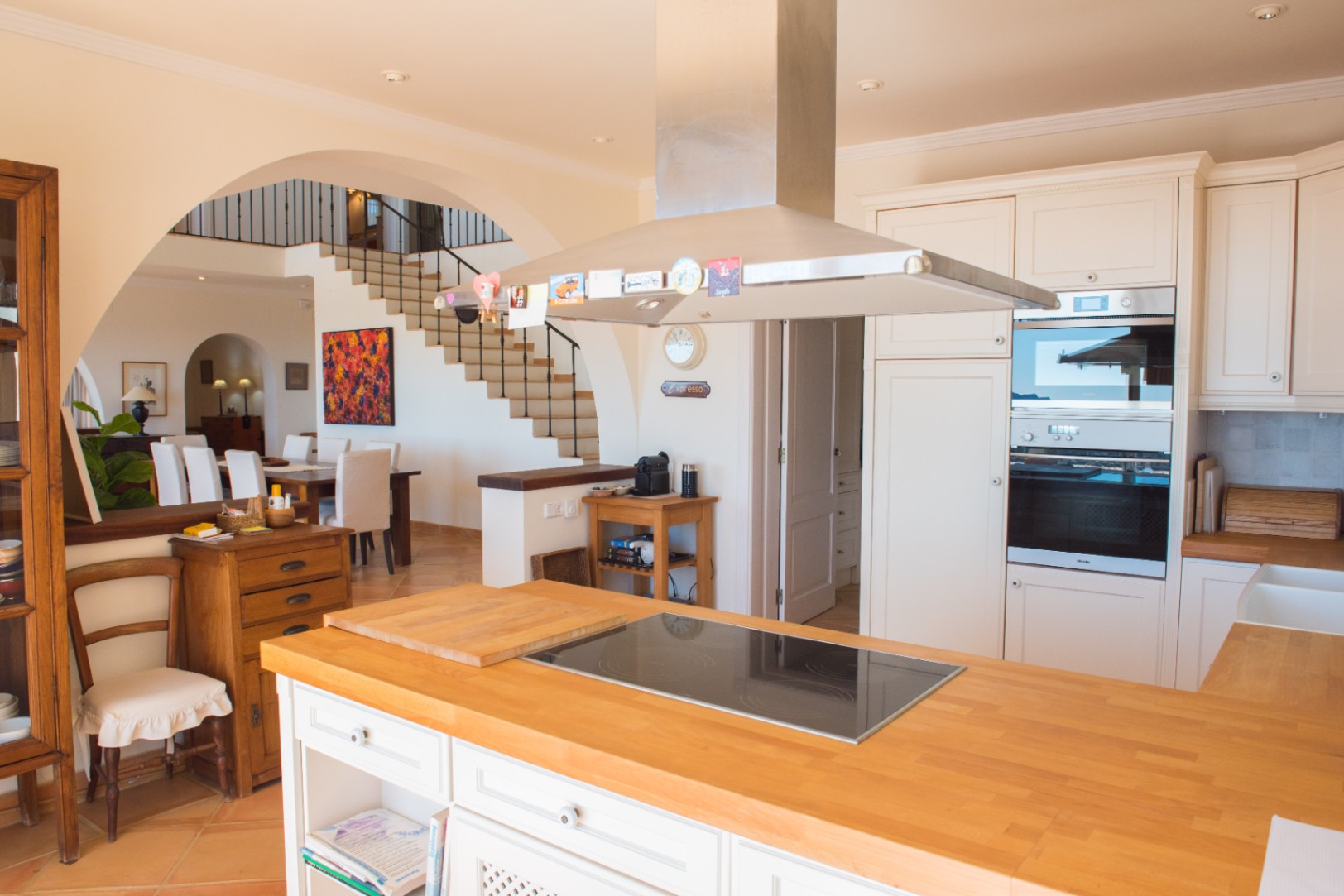 Open keuken met houten kookeiland, inductiekookplaat, roestvrijstalen afzuigkap en inbouwovens, grenzend aan de eetkamer en de trap met smeedijzer.