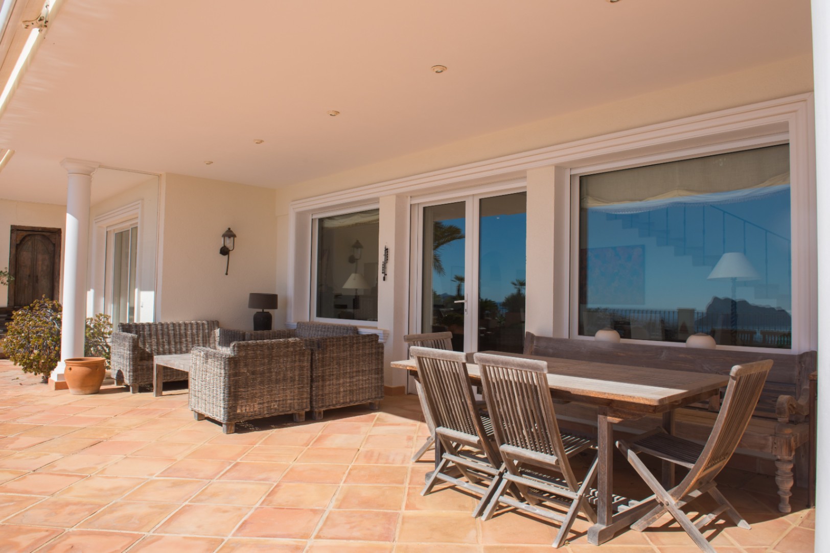 Overdekt terras met terracotta tegels, houten eethoek en wicker loungeset, grenzend aan grote glazen puien.