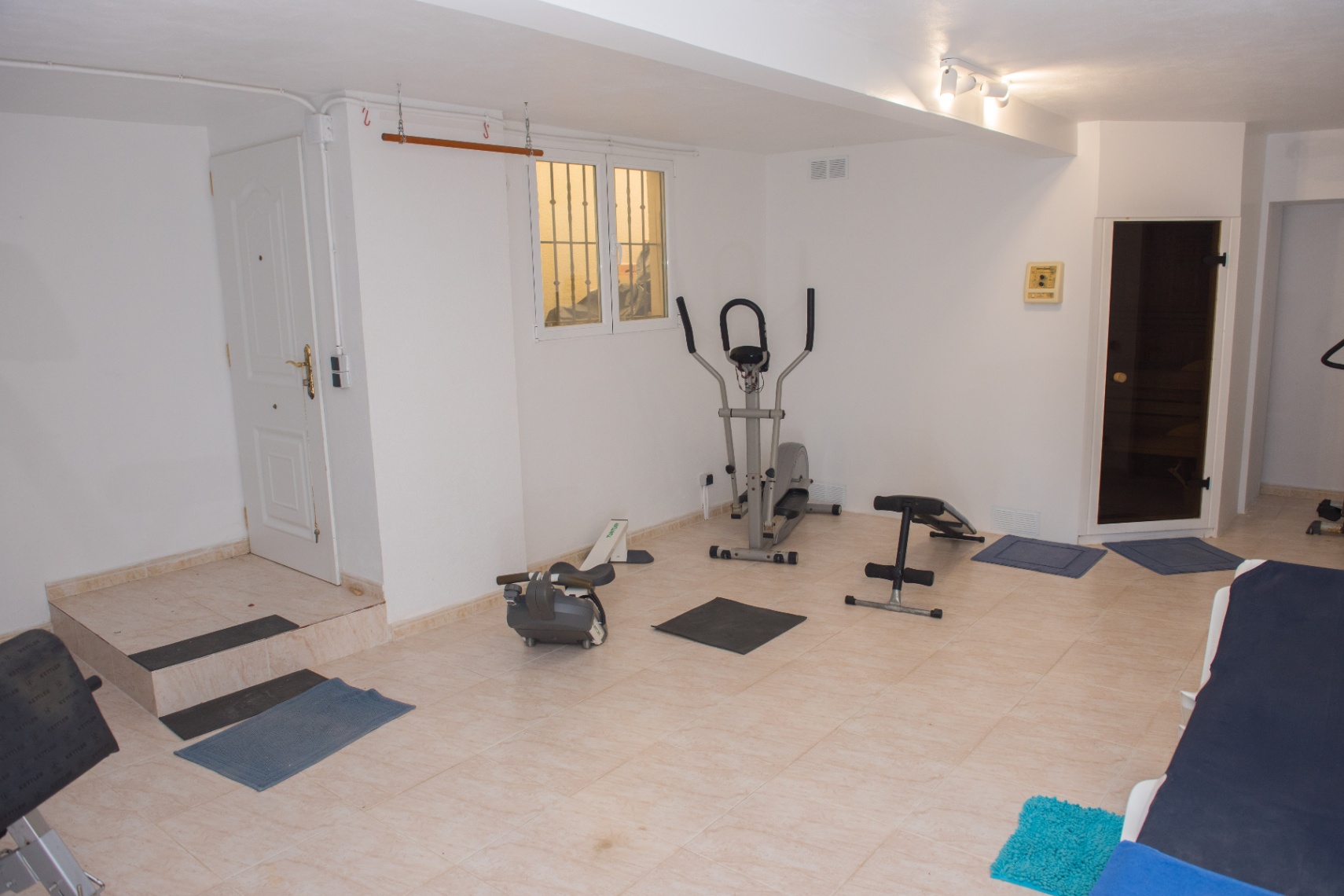 Privé fitnessruimte met cardio-apparatuur, een trainingsbank en directe toegang tot een sauna. De ruimte is afgewerkt met een lichte tegelvloer.