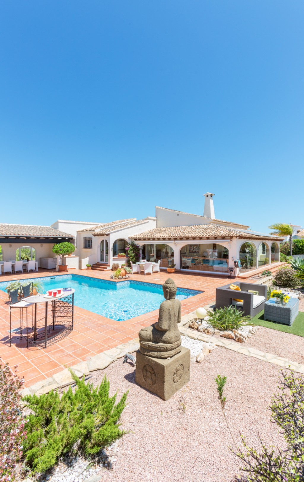 Buitenterras met zwembad, terracotta tegels, een stenen Boeddhabeeld en een mediterrane villa met een glazen veranda onder een pannendak.