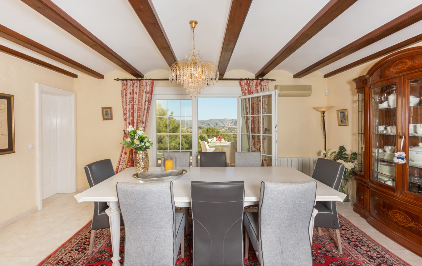 Eetkamer met houten balkenplafond, witte tafel en grijze stoelen. Voorzien van een klassieke servieskast en glazen deuren naar het terras met berguitzicht.