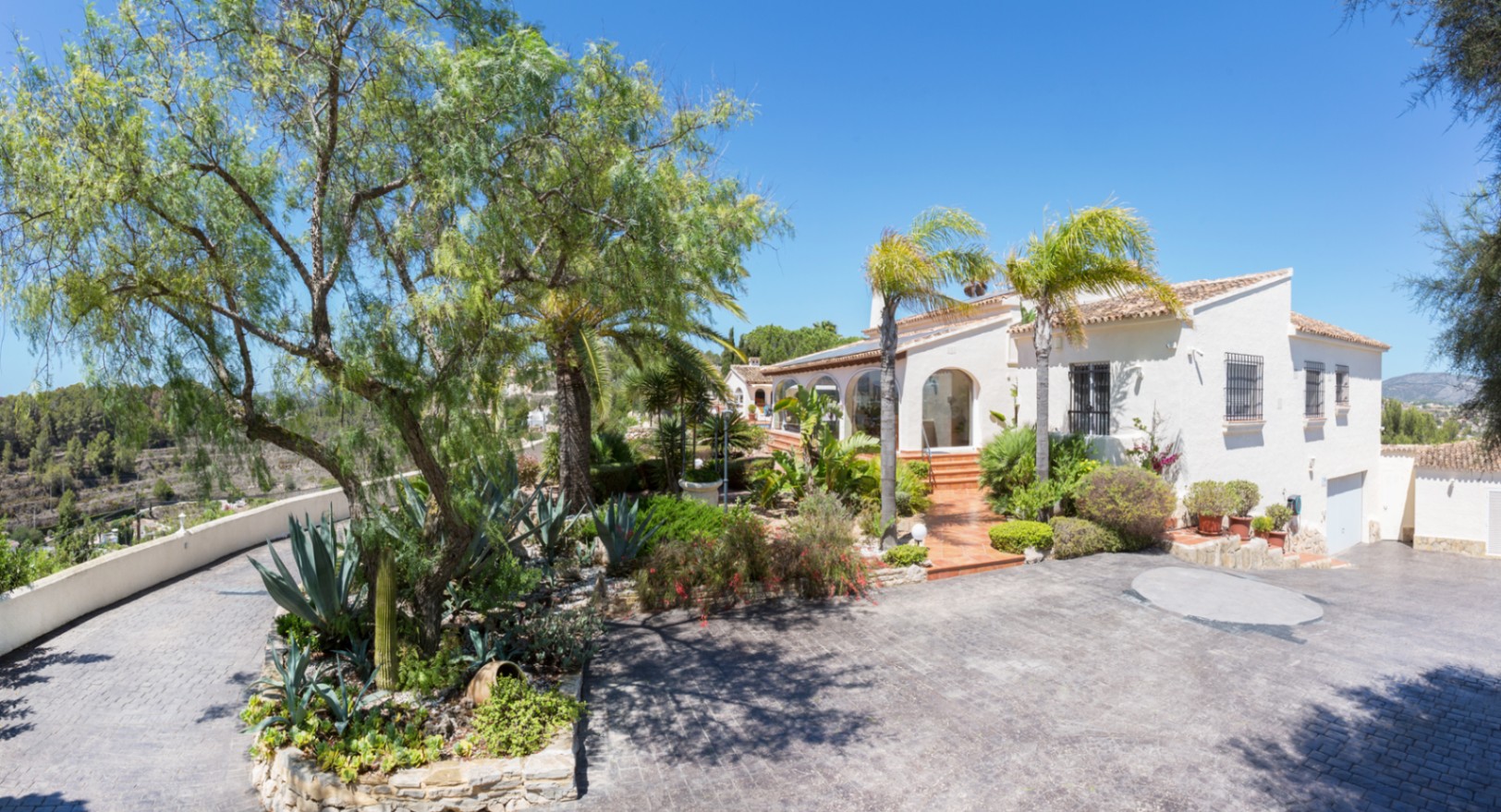 Exterieur van de mediterrane villa met een geplaveide oprit, aangelegde tuin met agaves en palmen, en een witte gevel met boogvormige architectuur.