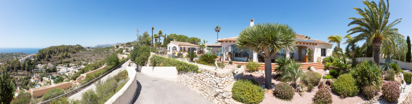 Panoramisch buitenaanzicht van de villa met een geplaveide oprit, natuurstenen muren en een mediterrane tuin met palmbomen en uitzicht op de kustlijn.