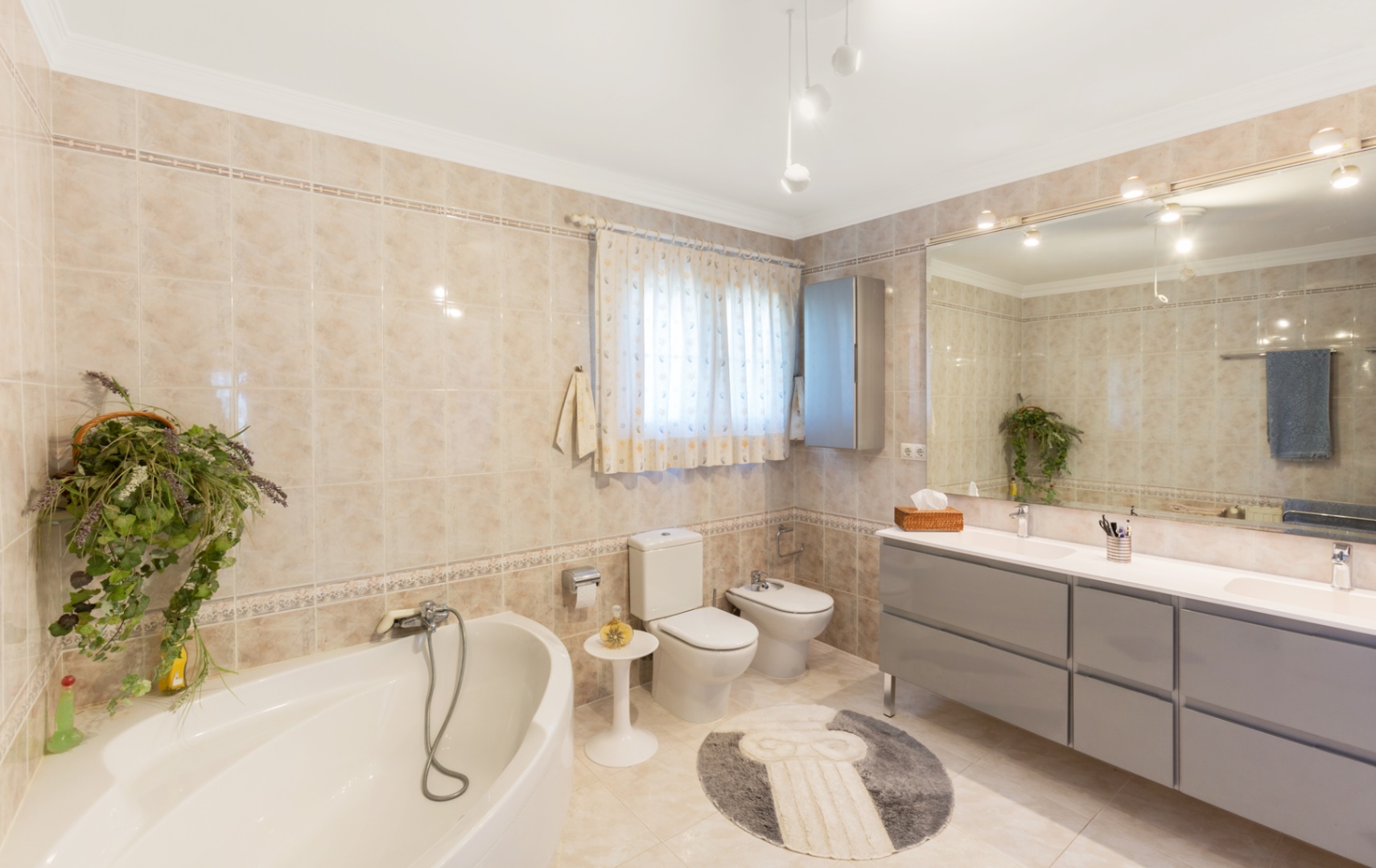 Ruime badkamer met hoekbad, dubbel wastafelmeubel in grijs en een grote wandspiegel. Beige betegeling met decoratieve sierranden op wanden en vloer.