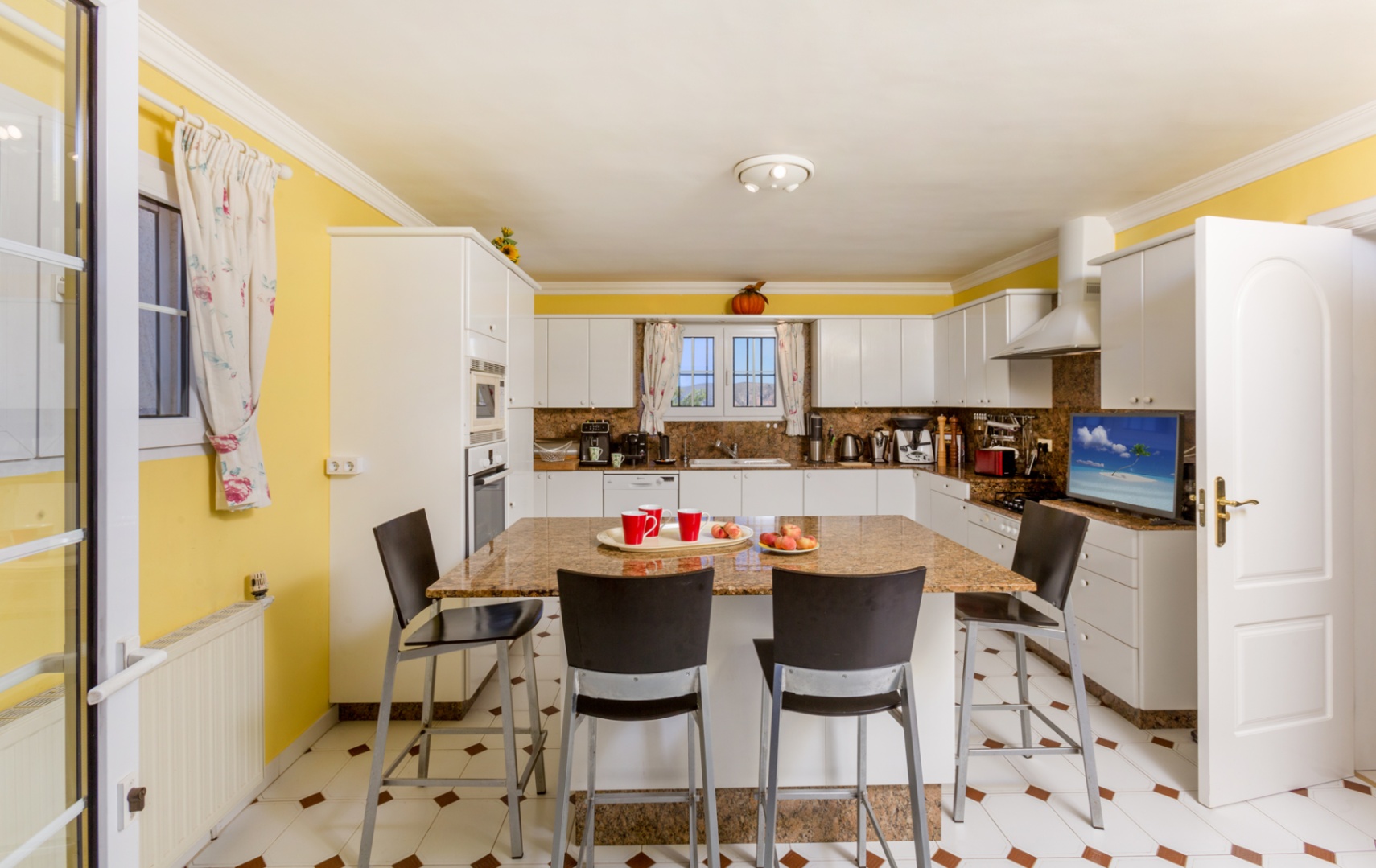 Ruime keuken met een granieten kookeiland, vier barkrukken, witte kasten en gele wanden. De tegelvloer is voorzien van bruine decoratieve accenten.