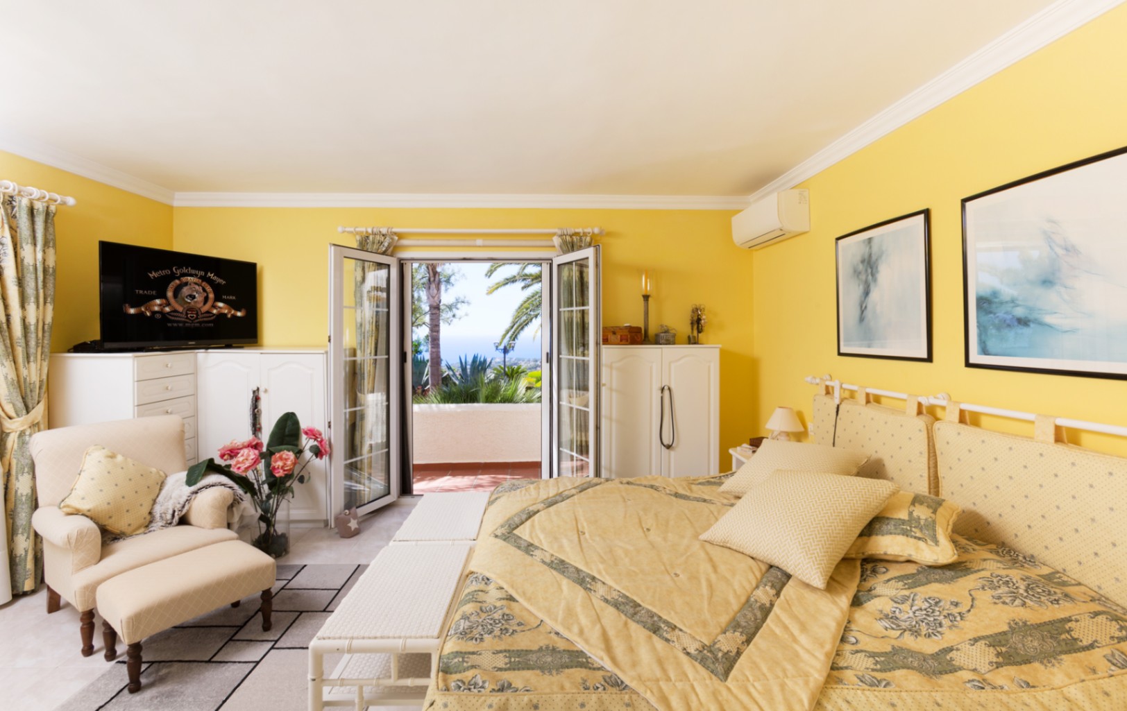 Slaapkamer met gele muren, witte kasten en een zithoek. Openslaande glazen deuren leiden naar een terras met uitzicht op zee, palmbomen en mediterrane planten.
