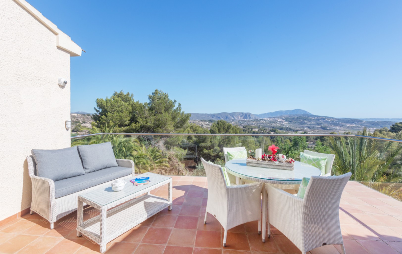 Terras met terracotta tegels, wit gevlochten tuinmeubilair en een glazen balustrade met uitzicht op de bergen en het omliggende mediterrane landschap.