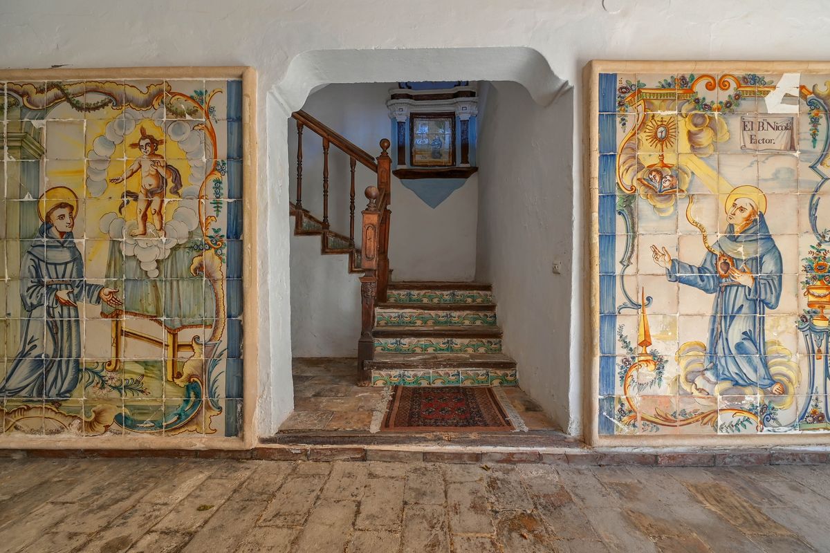 Entreehal met historische handbeschilderde keramische muurschilderingen, witgekalkte muren en een trap met gedecoreerde stootborden en houten leuning.