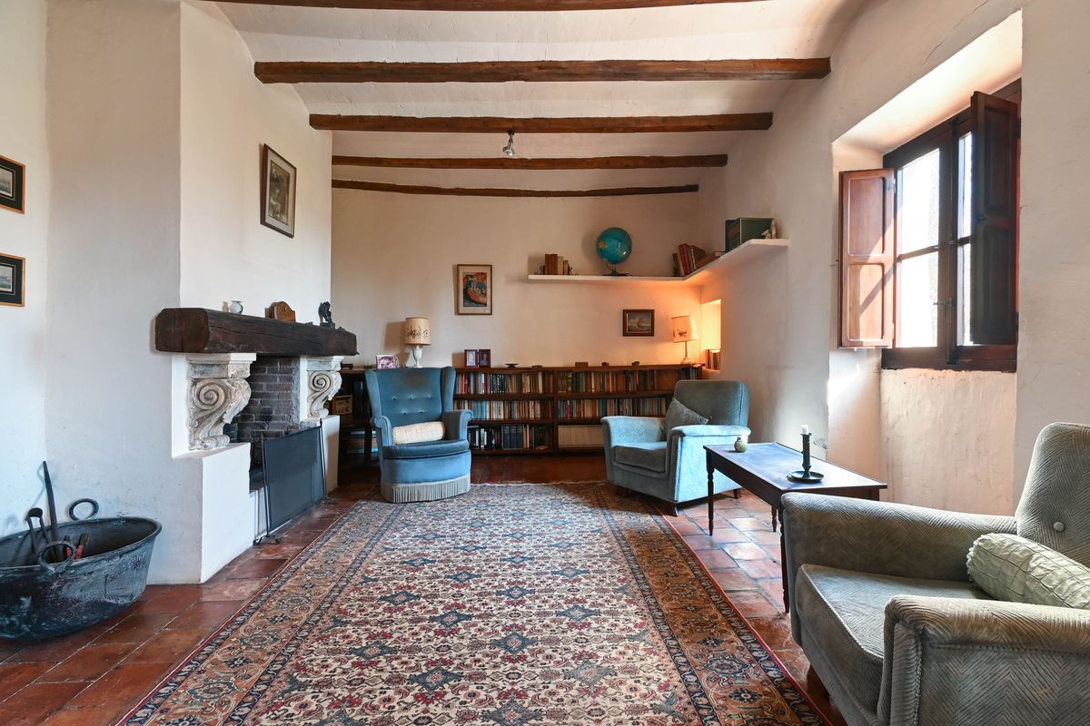 Historische woonkamer met houten balkenplafond, een grote open haard met houten latei, terracotta vloeren en boekenkasten.