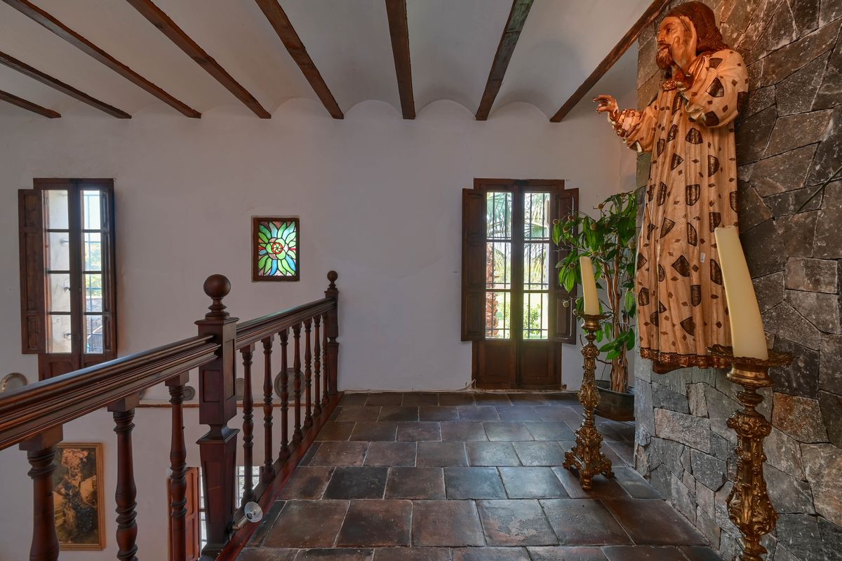 Overloop op de bovenverdieping met terracotta vloertegels, houten balkenplafond, een bewerkte balustrade en een natuurstenen muur met religieuze decoratie.