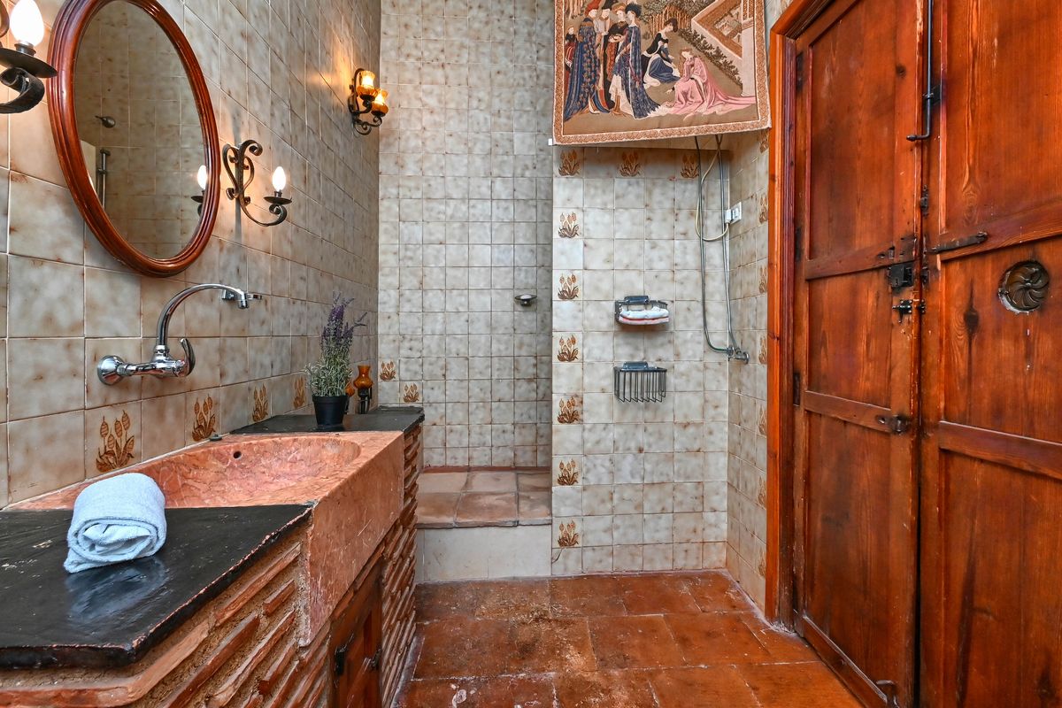 Rustieke badkamer met een roze natuurstenen wastafel, gedecoreerde wandtegels, smeedijzeren wandlampen en een authentieke massief houten deur.