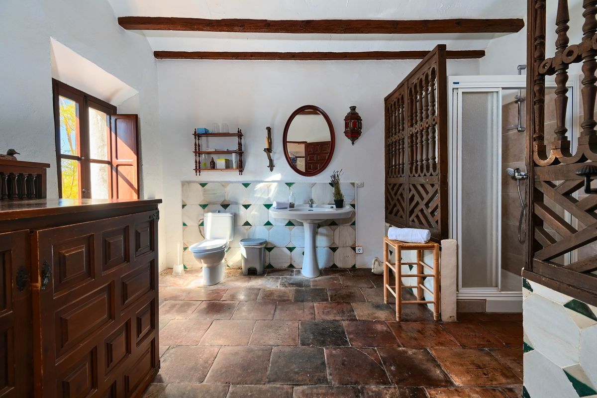 Rustieke badkamer met terracotta vloeren, houten balkenplafond en een gesneden houten scherm. Voorzien van een wastafel op zuil en decoratieve wandtegels.