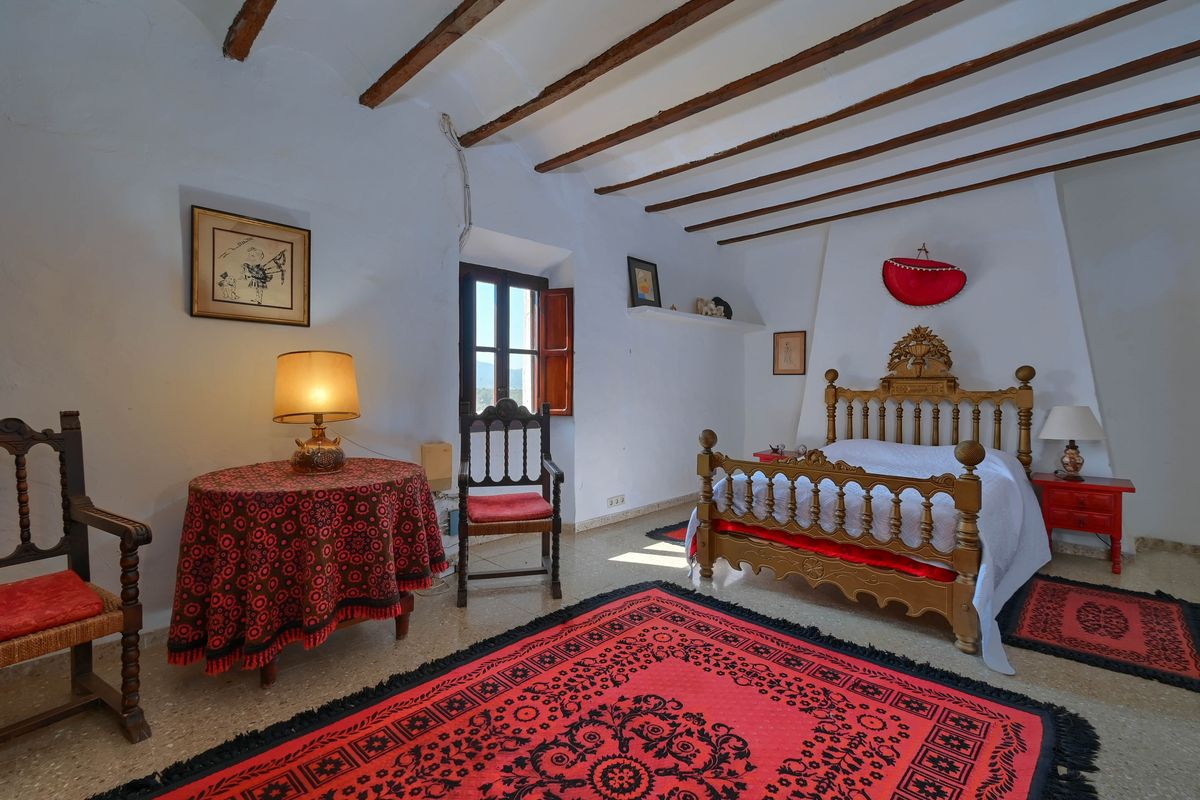 Rustieke slaapkamer met originele houten plafondbalken, een handgesneden bedframe en decoratieve rode tapijten op een stenen vloer.