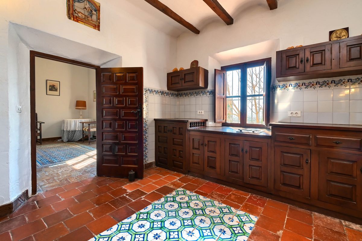 Traditionele keuken met donkerhouten kasten, terracotta vloer met decoratieve tegels en zichtbare balken. Een zware houten deur biedt toegang tot de aangrenzende kamer.