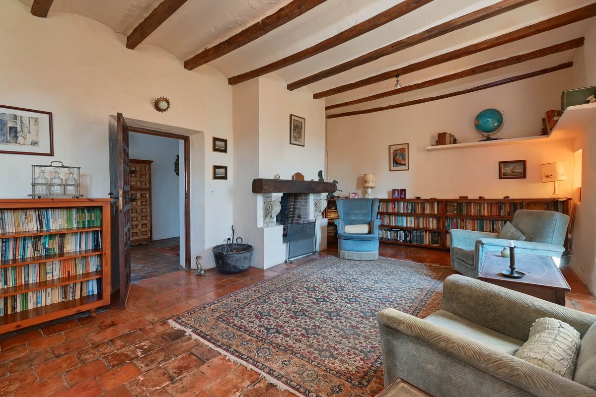 Woonkamer met authentieke houten plafondbalken, een klassieke haard met houten balk, terracotta vloer en wandvullende boekenkasten.