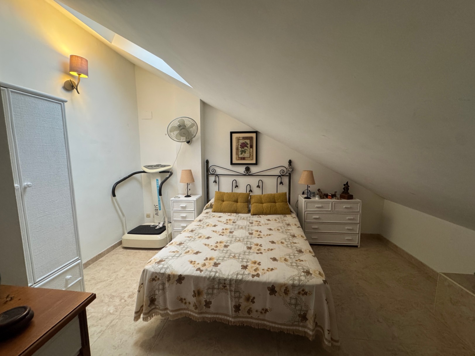 Zolderkamer met tweepersoonsbed, fitnessapparaat en witte kasten.