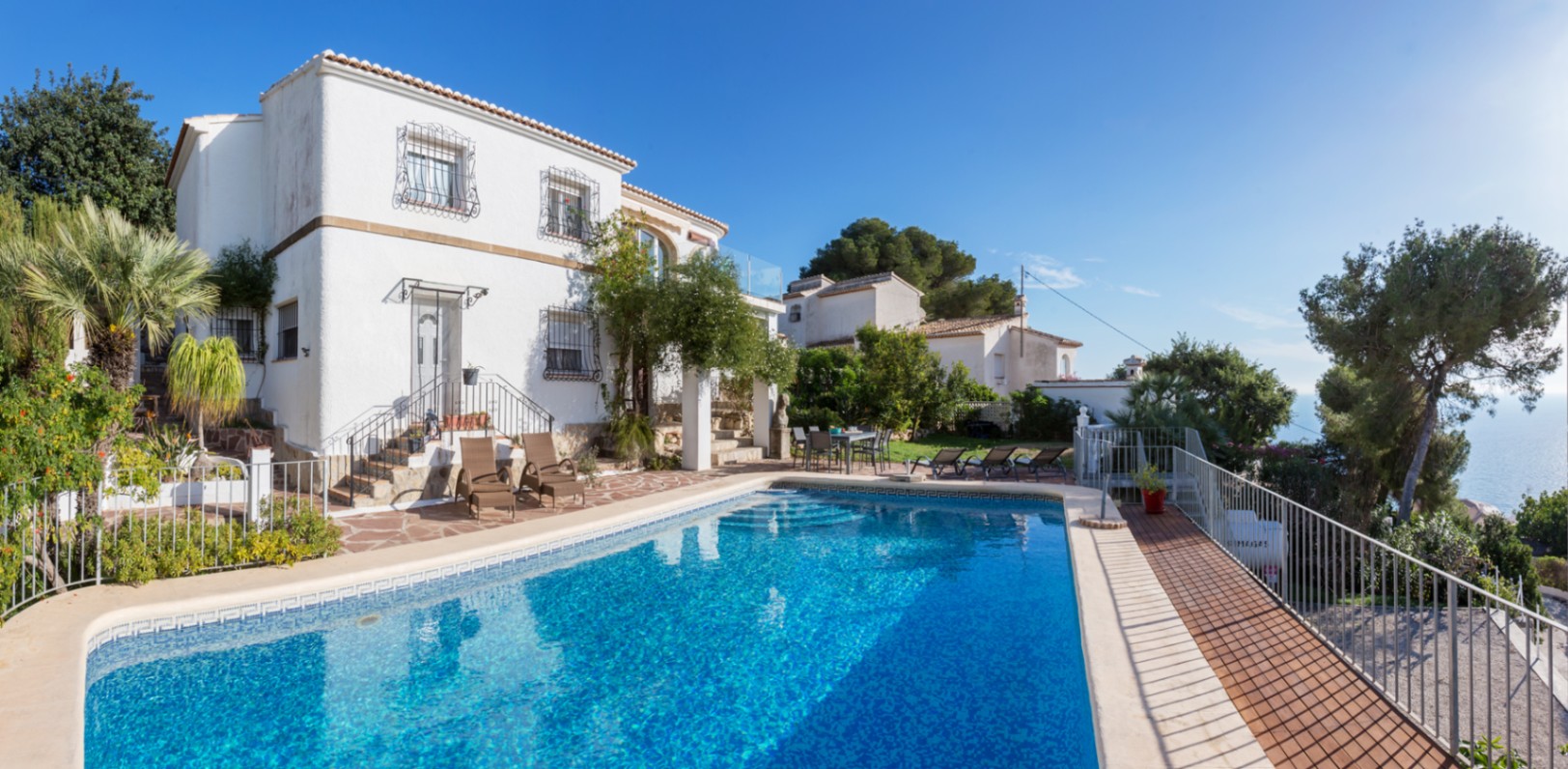 Exterieur van de mediterrane villa met groot zwembad, natuurstenen terras met ligbedden en witte gevel met traditioneel smeedwerk en uitzicht op zee.