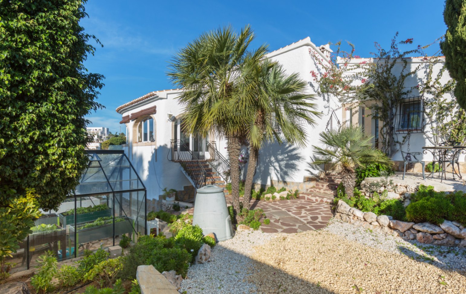 Exterieur van een witte mediterrane villa met natuurstenen paden, palmbomen en een glazen plantenkas in een verzorgde, aangelegde tuin.