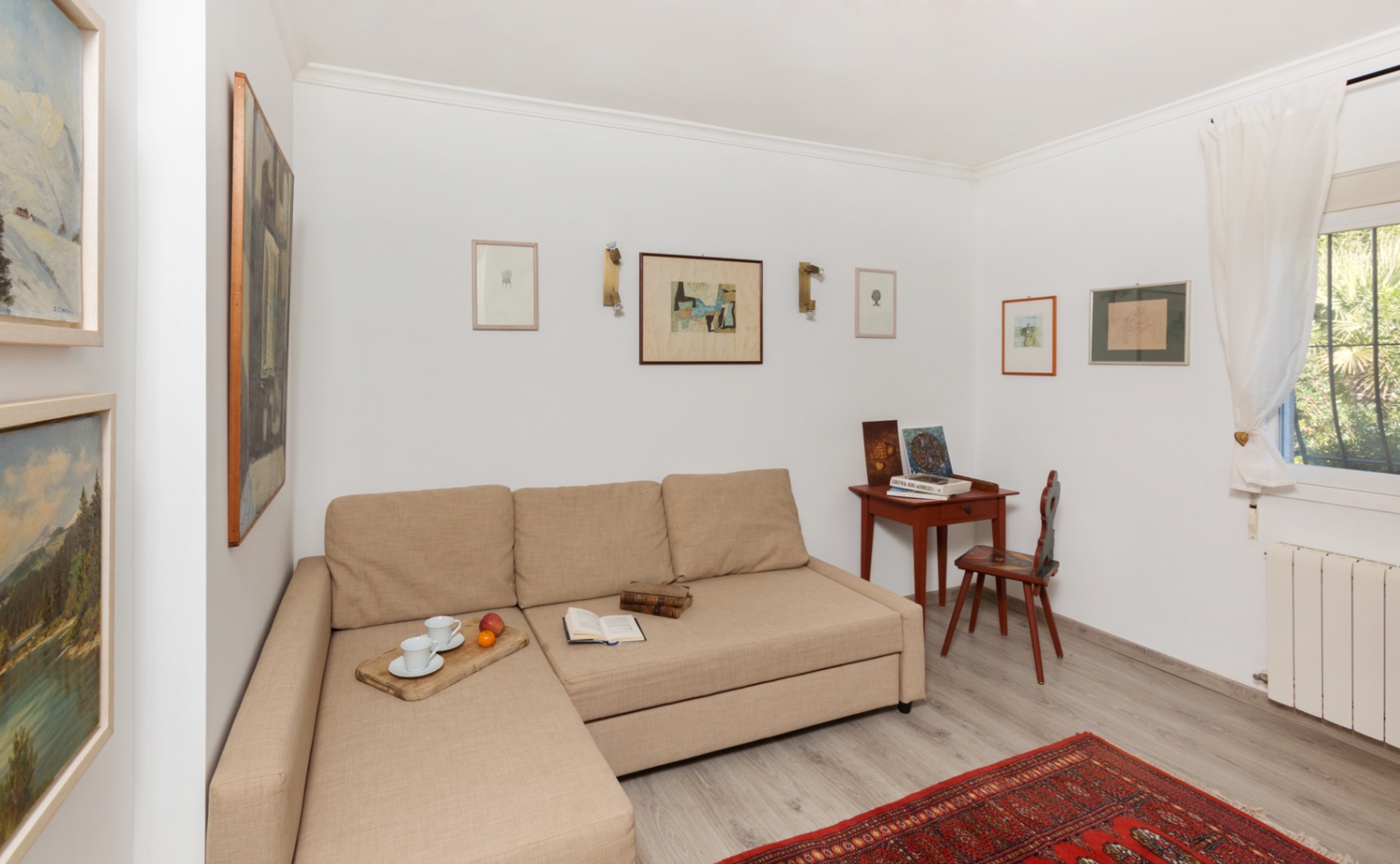 Lichte zithoek met een beige chaise longue bank, een klassiek houten bureau en een wand met ingelijste kunstwerken op een grijze laminaatvloer.