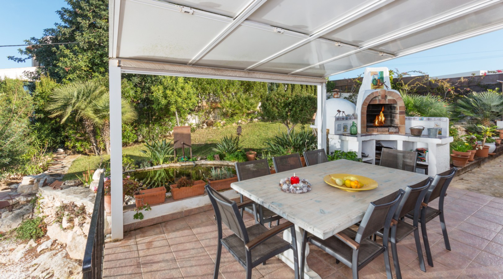 Overdekt terras met grote houten eettafel, traditionele stenen houtoven en uitzicht op de mediterrane tuin met vijver.