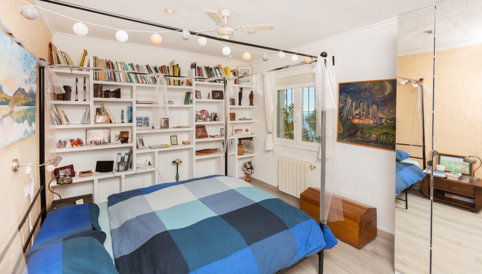 Slaapkamer met zwart metalen hemelbed, ingebouwde witte boekenkast en een grote spiegelwand. Voorzien van een plafondventilator en raam met uitzicht op zee.