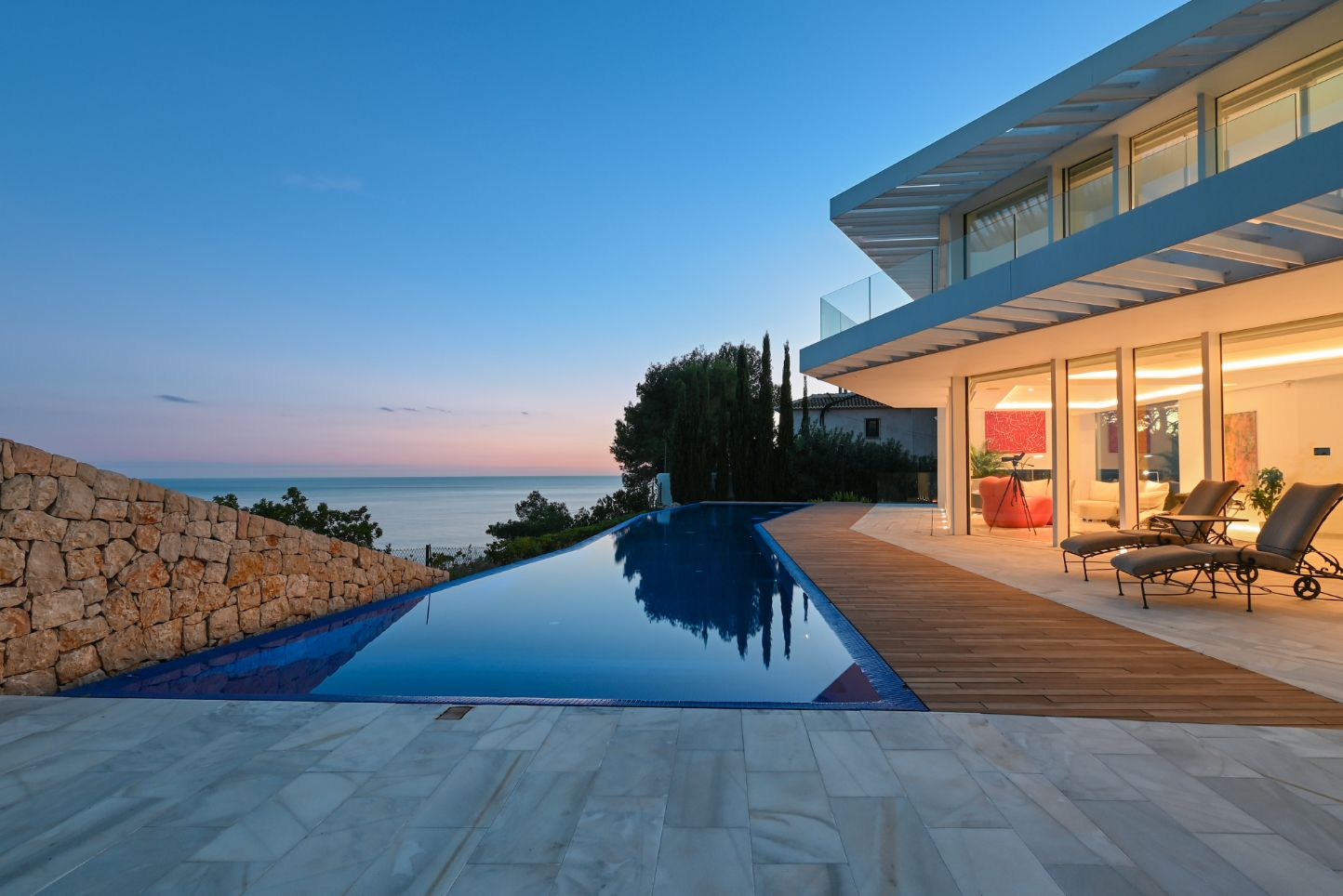 Infinity zwembad met houten terras en stenen bestrating met uitzicht op zee. Moderne villa met grote glaspartijen en natuurstenen muur bij zonsondergang.