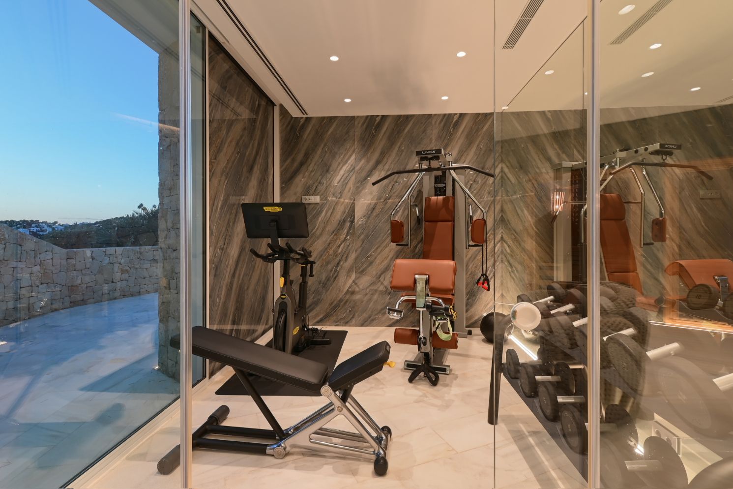Moderne fitnessruimte met Technogym-apparatuur, wanden van geaderd natuursteen en glazen wanden die uitkijken op een terras van natuursteen.