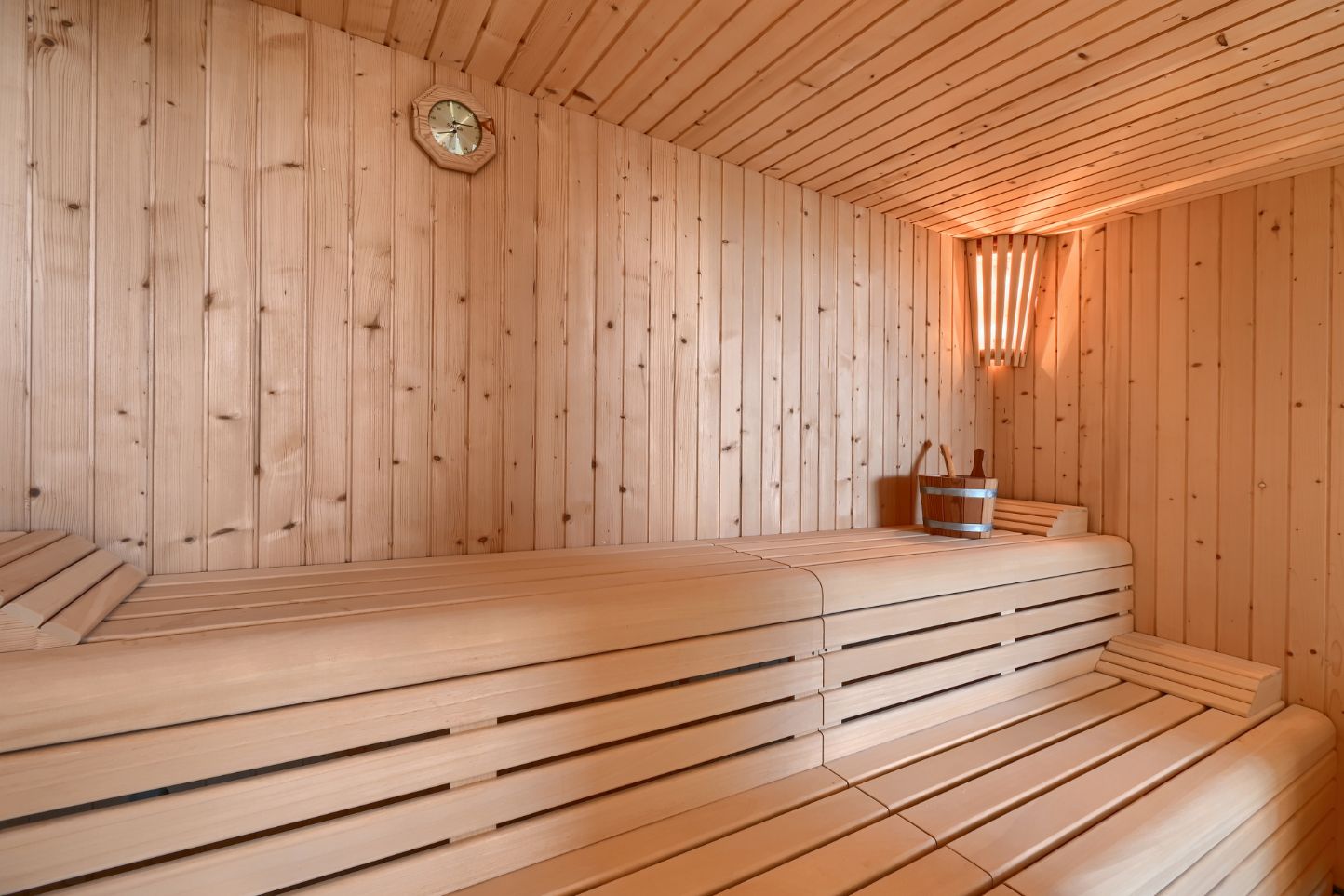 Sauna-interieur met lichte houten afwerking, gelaagde lattenbanken, hoekverlichting en traditionele houten accessoires zoals een emmer en pollepel.
