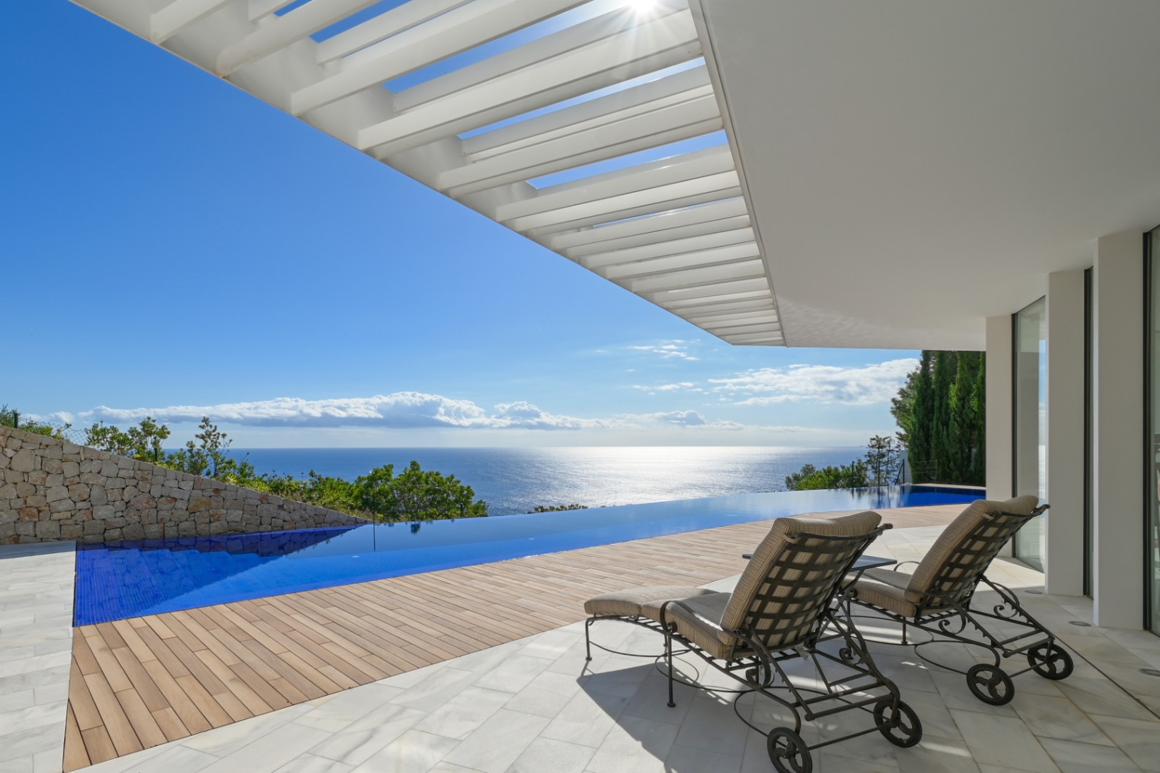 Terras met overloopzwembad en panoramisch uitzicht op zee, houten vlonders en marmeren vloer onder een witte pergola met ligbedden.