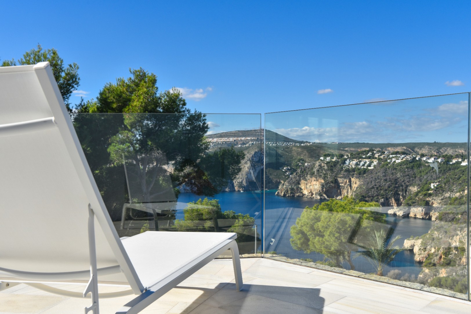 Terras met witte ligstoel en glazen balustrade, uitkijkend over een mediterrane baai en rotsachtige kustlijn met pijnbomen.