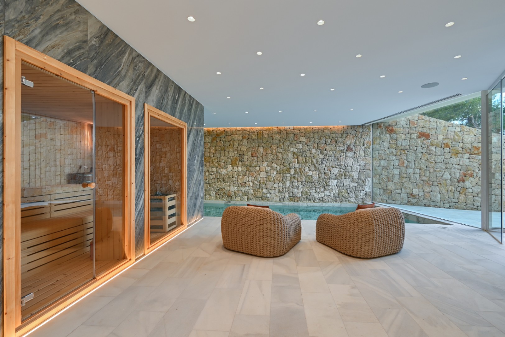 Wellnessruimte binnen met houten sauna, gevlochten loungestoelen en een natuurstenen accentmuur grenzend aan het binnenzwembad.
