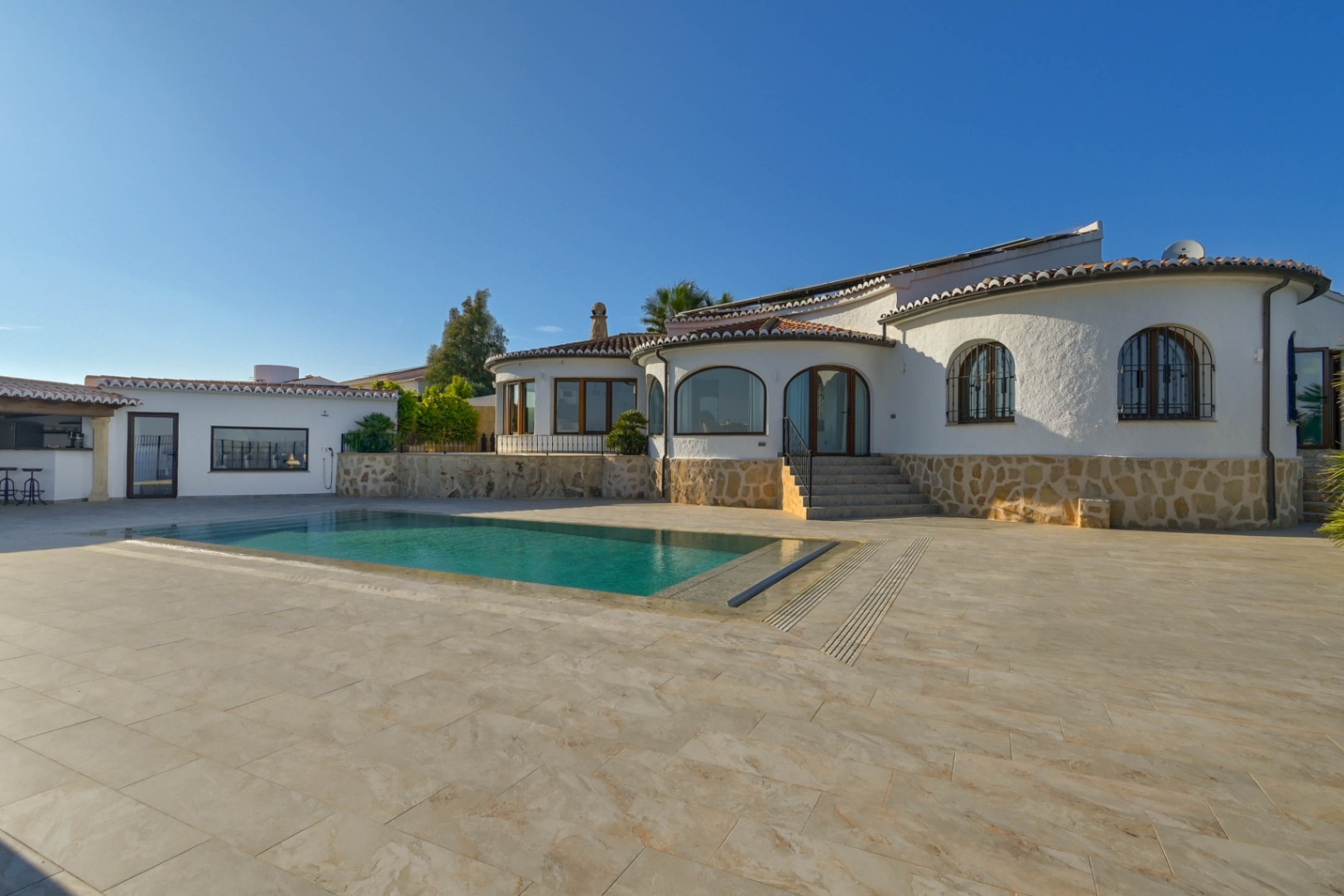 Exterieur van een mediterrane villa met rechthoekig zwembad, natuurstenen plint en ruim betegeld terras onder een heldere hemel.