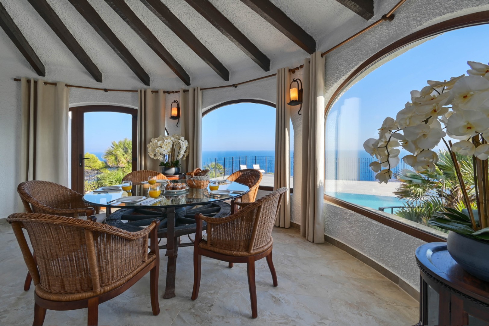 Ronde eetkamer met een glazen tafel op een wagenwiel en rotan stoelen, onder een balkenplafond met panoramisch uitzicht op zee door de karakteristieke boogramen.