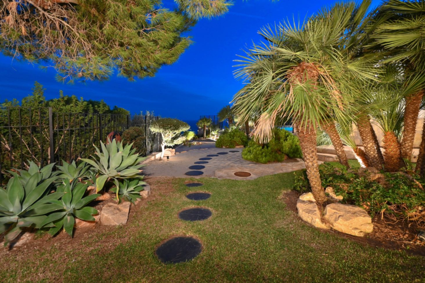 Tuinpad met ronde stapstenen in een mediterrane tuin met palmbomen, agaves en geïntegreerde buitenverlichting tijdens de avondschemering.