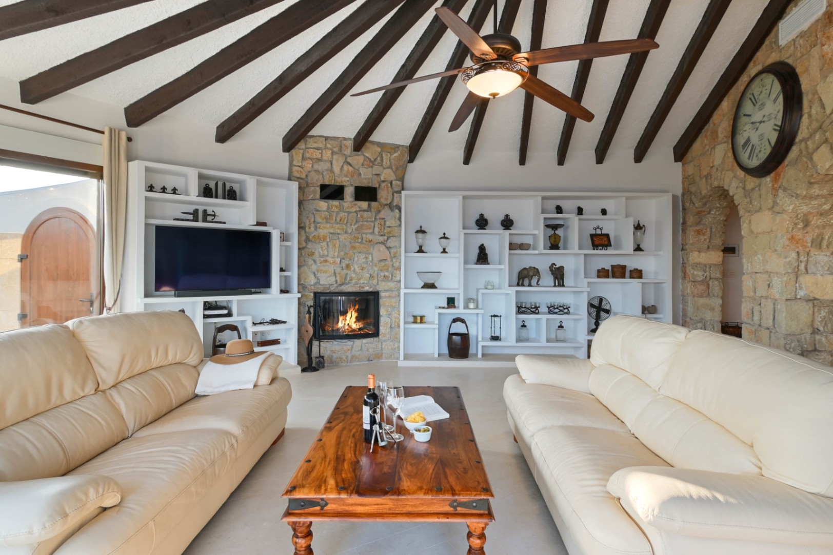 Woonkamer met een balkenplafond, natuurstenen open haard en ingebouwde witte kasten. Voorzien van crèmekleurige lederen banken en een rustieke houten salontafel.