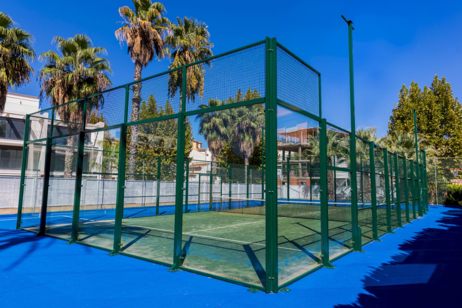 Buiten padelbaan met groen kunstgras, wanden van gehard glas en een groen metalen frame, omgeven door palmbomen en een blauwe ondergrond.