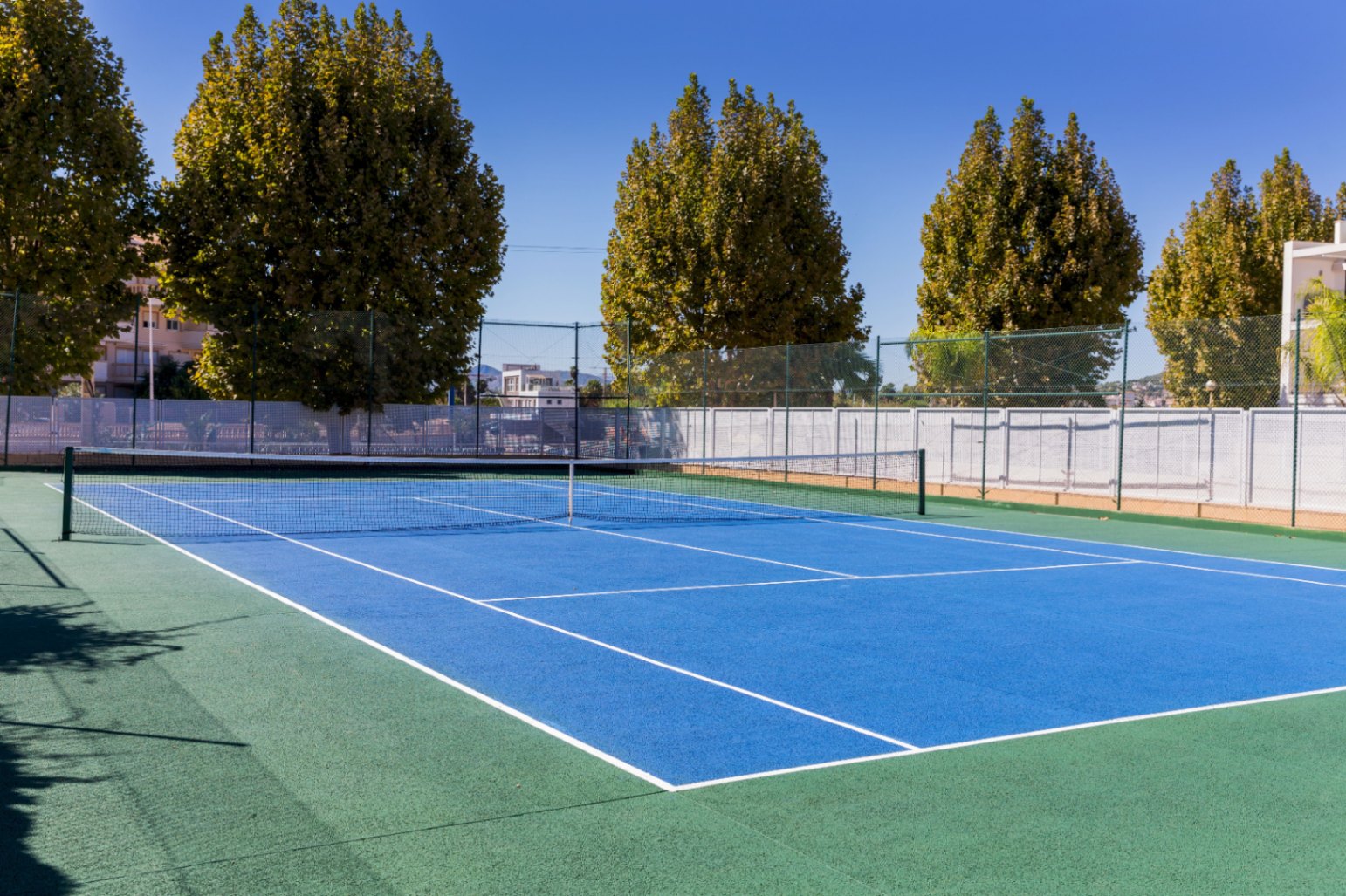 Buiten tennisbaan met een harde ondergrond, blauw speelveld en groene uitloopstrook, omheind met gaashekwerk en omgeven door volwassen bomen.