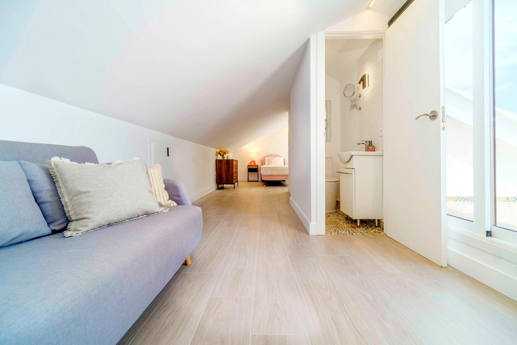 Lichte zolderkamer met schuine witte plafonds, houtlook vloertegels, een zithoek en een aangrenzende badkamer met decoratieve patroontegels.