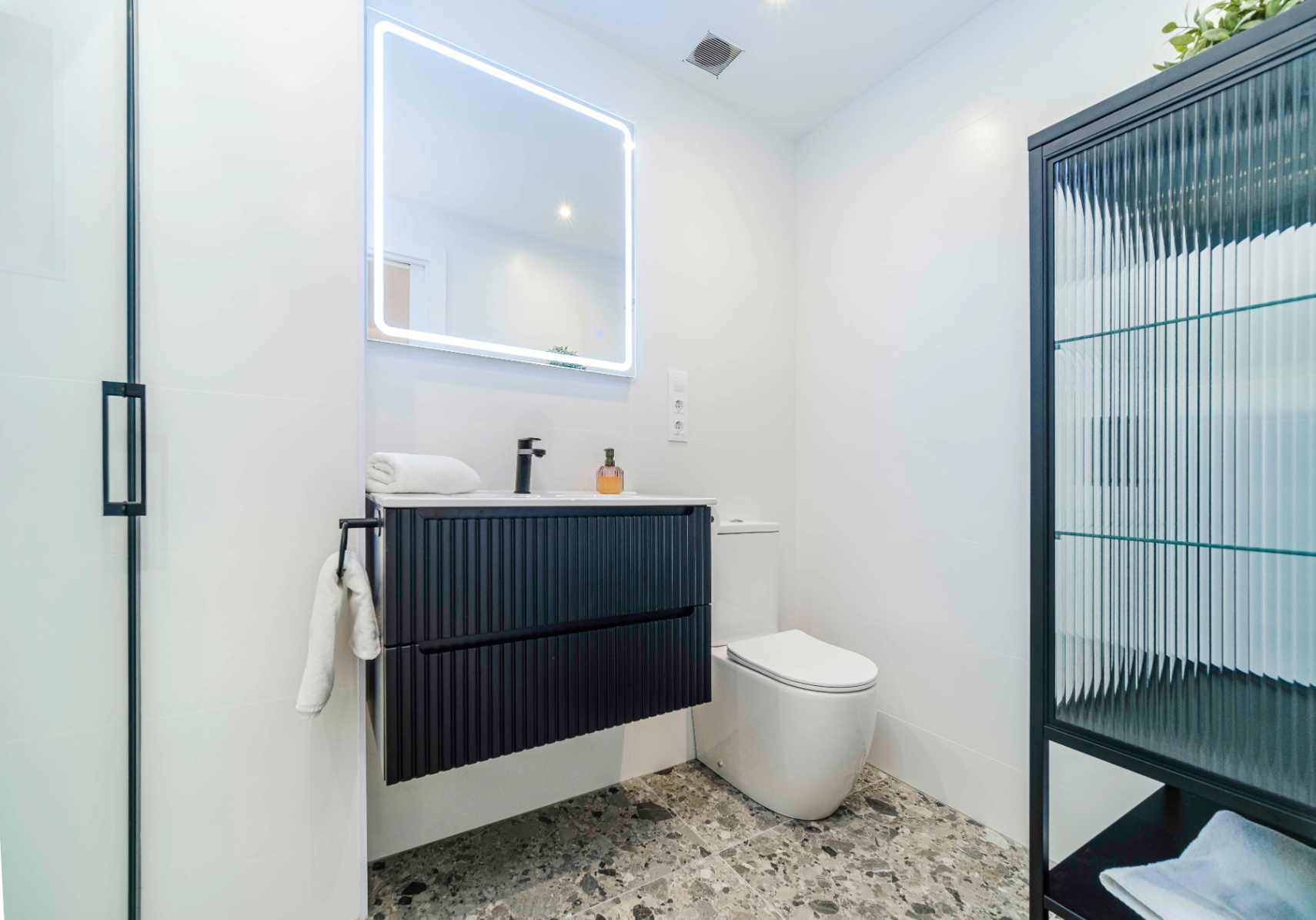 Moderne badkamer met een zwart geribbeld zwevend wastafelmeubel, LED-spiegel en een grijze terrazzovloer.