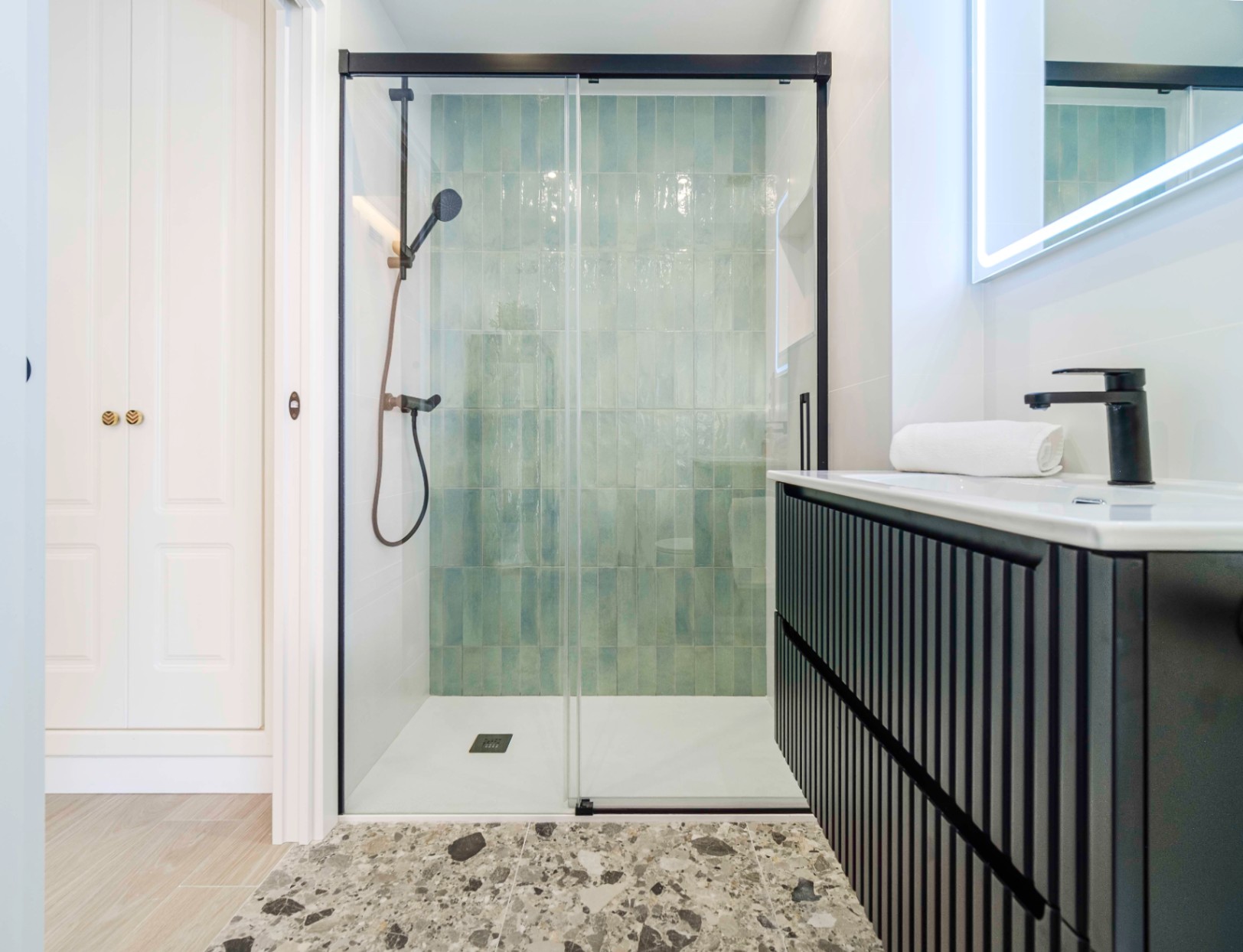 Moderne badkamer met inloopdouche voorzien van turquoise verticale tegels, zwarte kranen en een zwart geribbeld wastafelmeubel met verlichte spiegel.