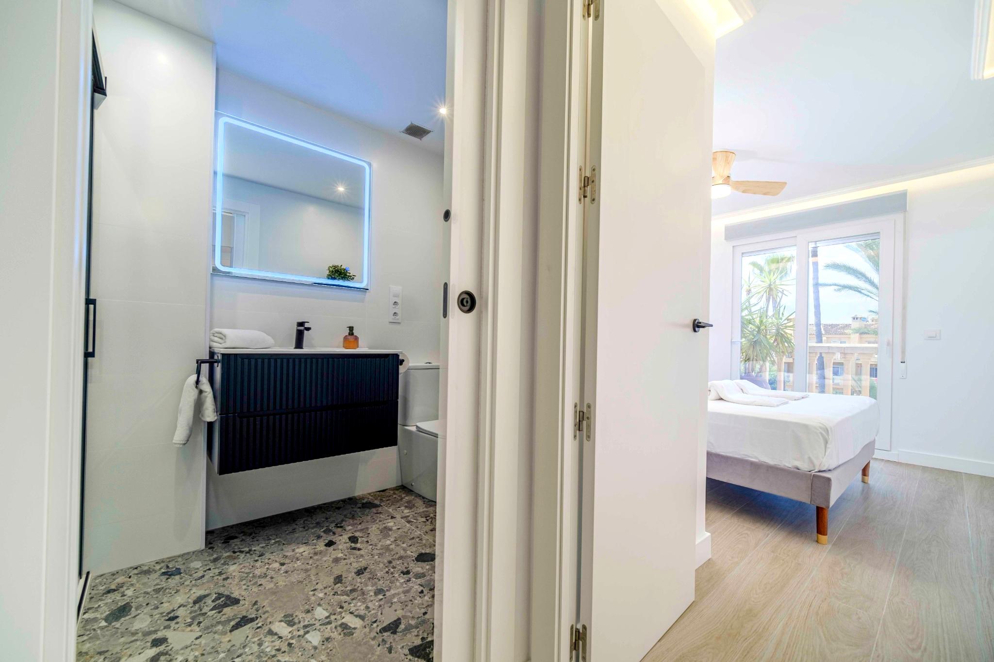 Moderne badkamer met terrazzovloer en zwart meubel, grenzend aan een lichte slaapkamer met balkon en uitzicht op de groene omgeving.