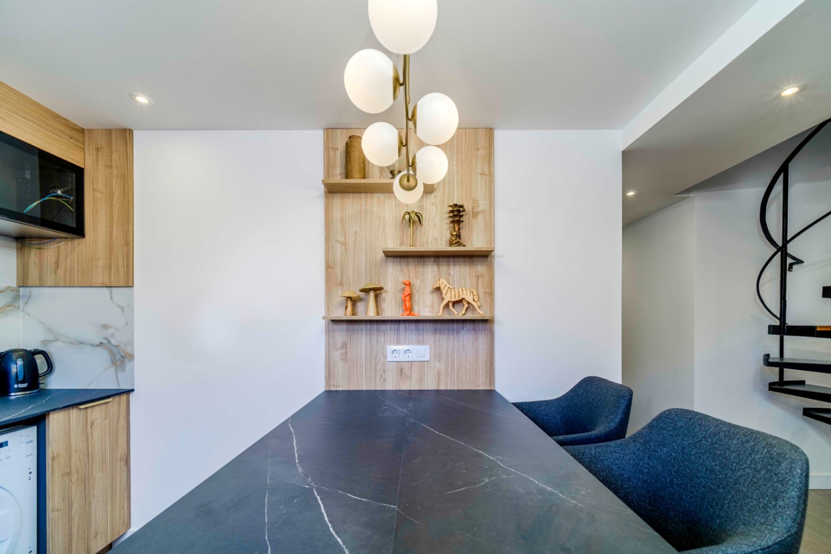 Moderne eethoek met een tafel in donker marmerlook, blauwe gestoffeerde stoelen en houten wandplanken, verlicht door een design bolkroonluchter.