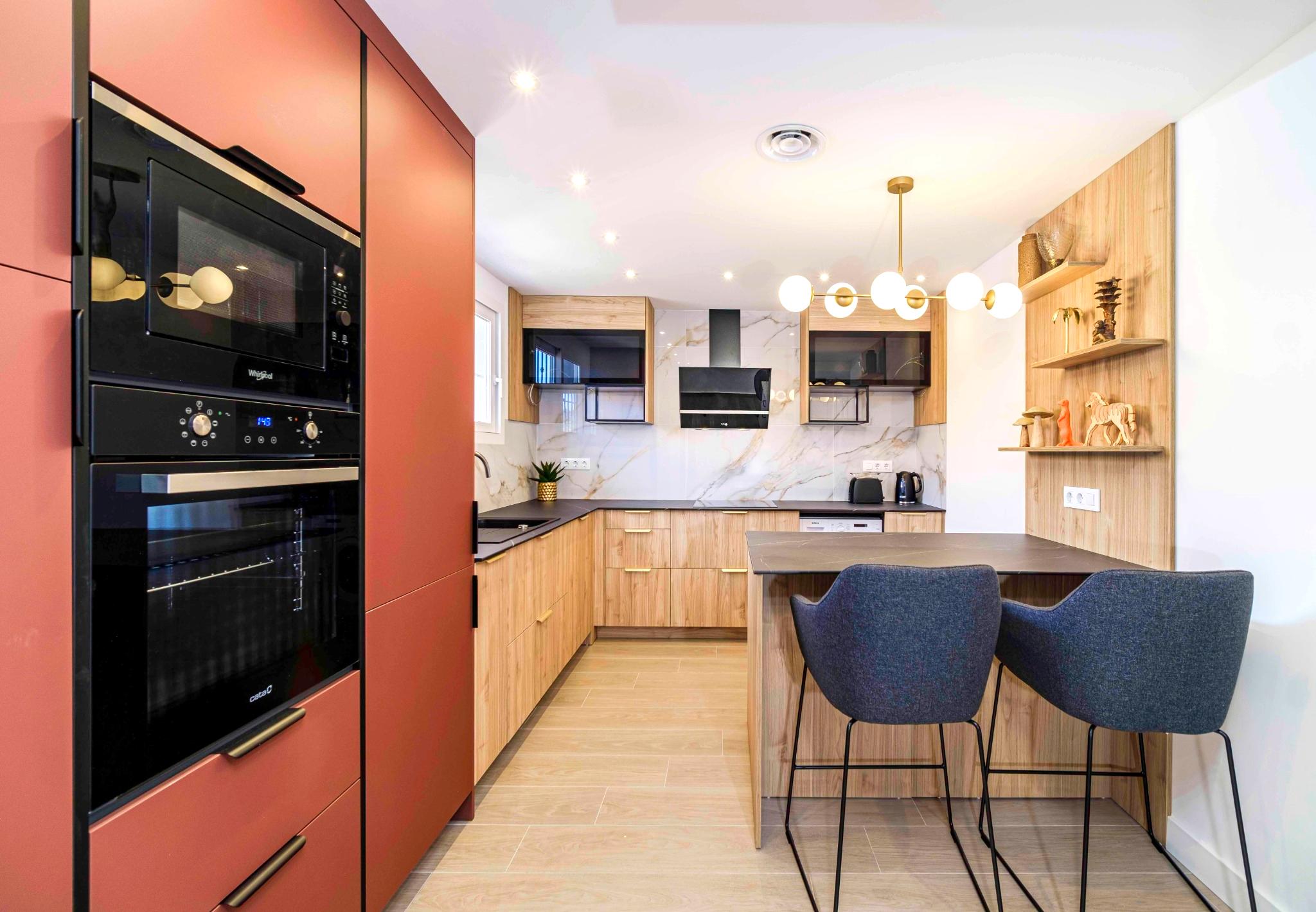 Moderne keuken met terracotta kastenwand, licht houten onderkasten, marmerlook achterwand en een kookeiland met blauwe barkrukken en een gouden bollamp.