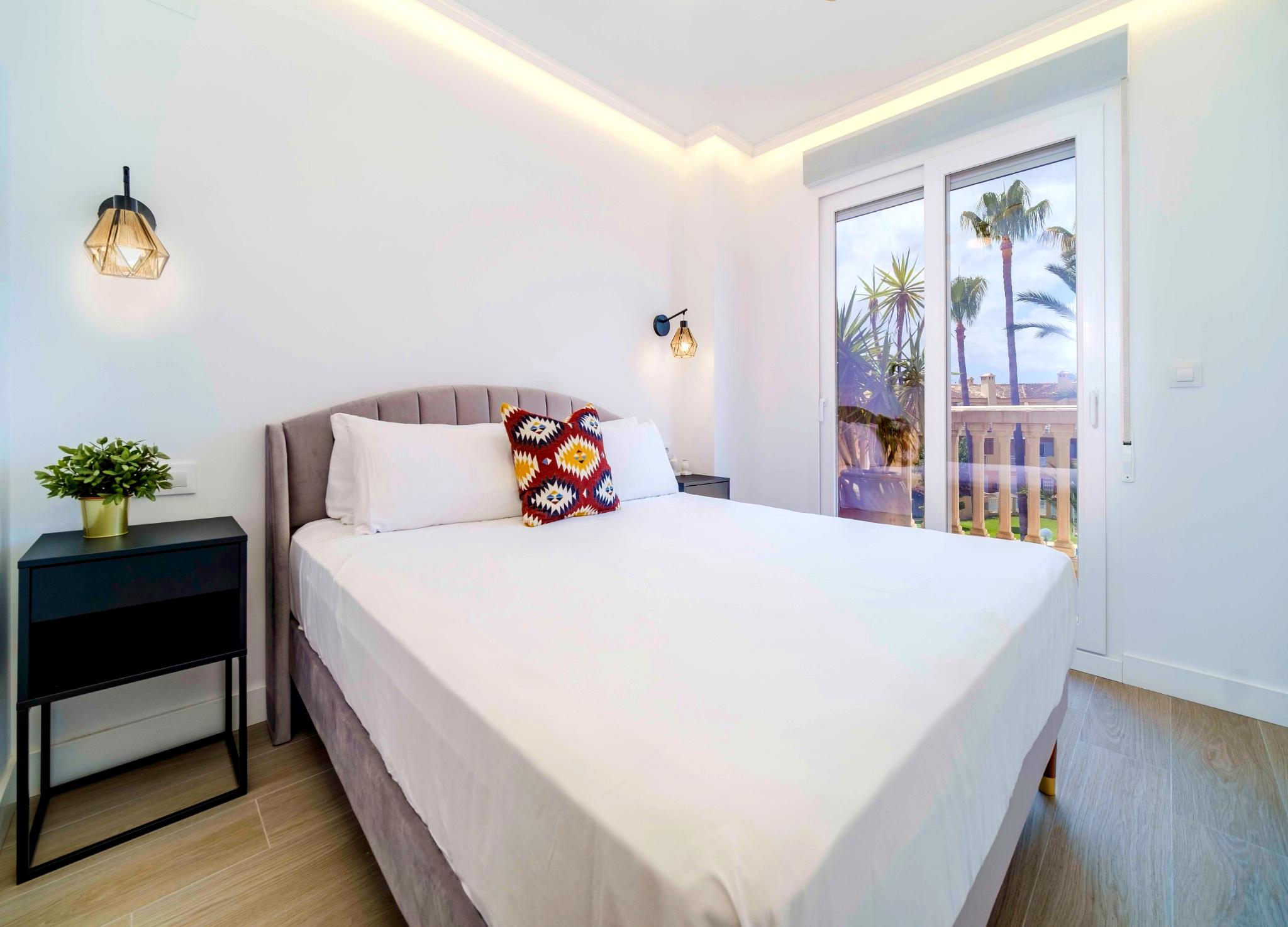 Moderne slaapkamer met fluwelen hoofdbord, geometrische wandlampen en indirecte plafondverlichting. Grote schuifdeuren bieden toegang tot een balkon met uitzicht op palmbomen.