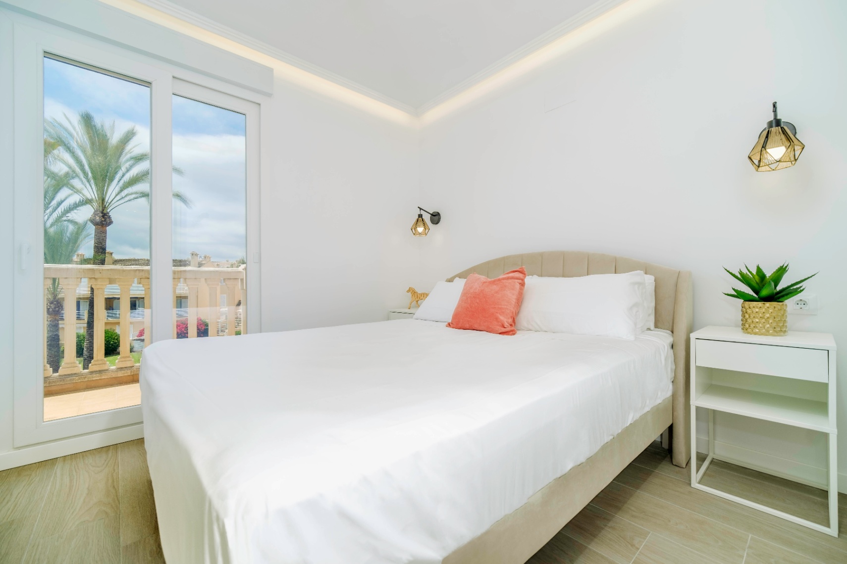 Moderne slaapkamer met gestoffeerd hoofdbord, geometrische wandlampen en een glazen schuifdeur die toegang biedt tot een terras met uitzicht op palmbomen.