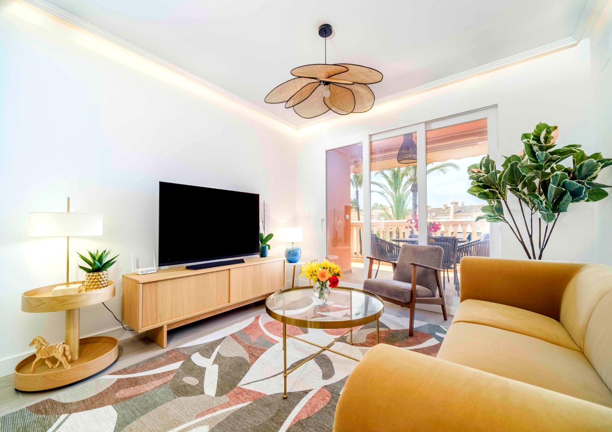 Moderne woonkamer met gele bank, een bloemvormige hanglamp en glazen deuren die toegang bieden tot een terras met uitzicht op palmbomen.