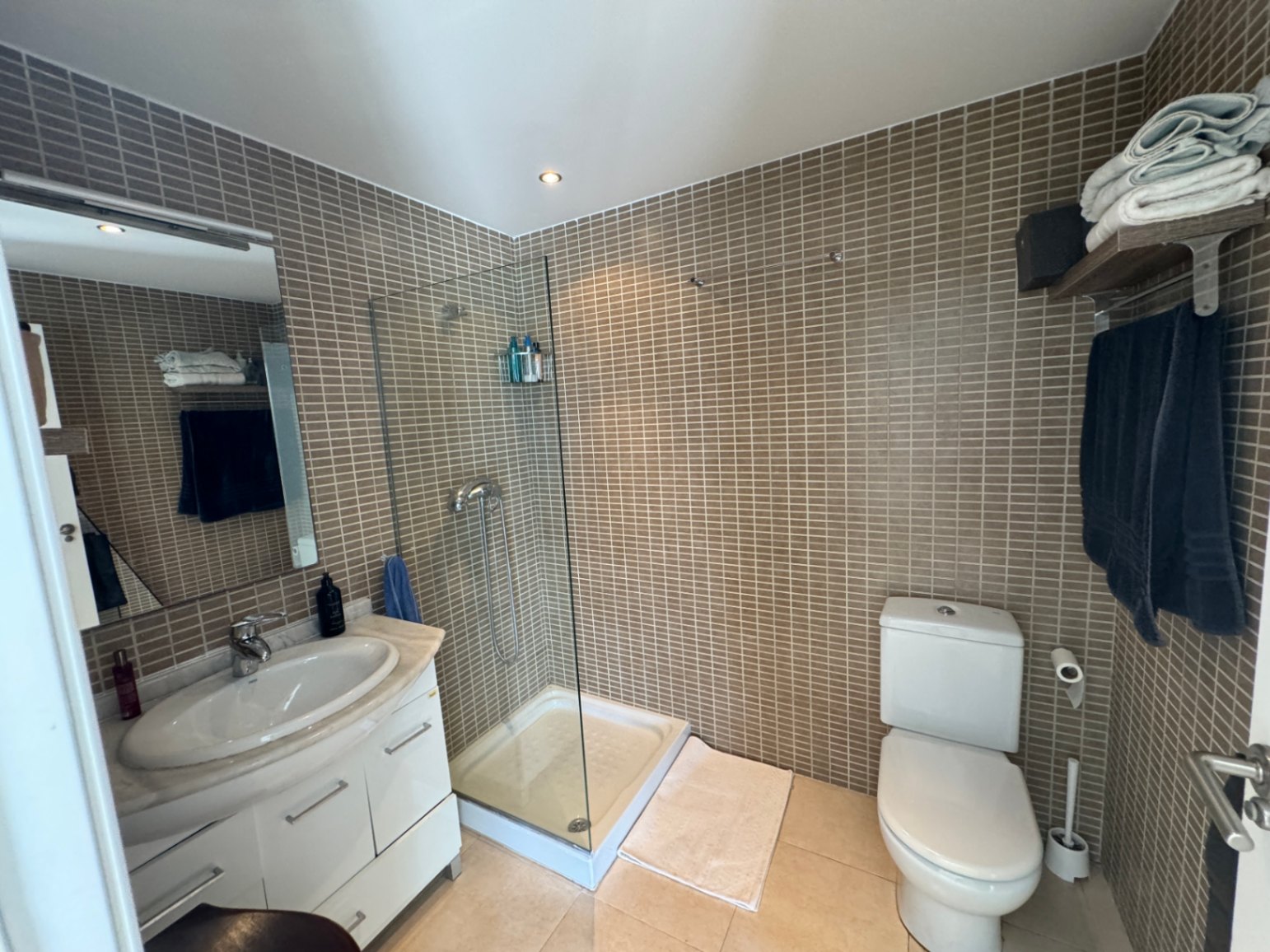 Badkamer met glazen inloopdouche, bruine mozaïektegels en een wit wastafelmeubel met marmeren blad en inbouwspots.