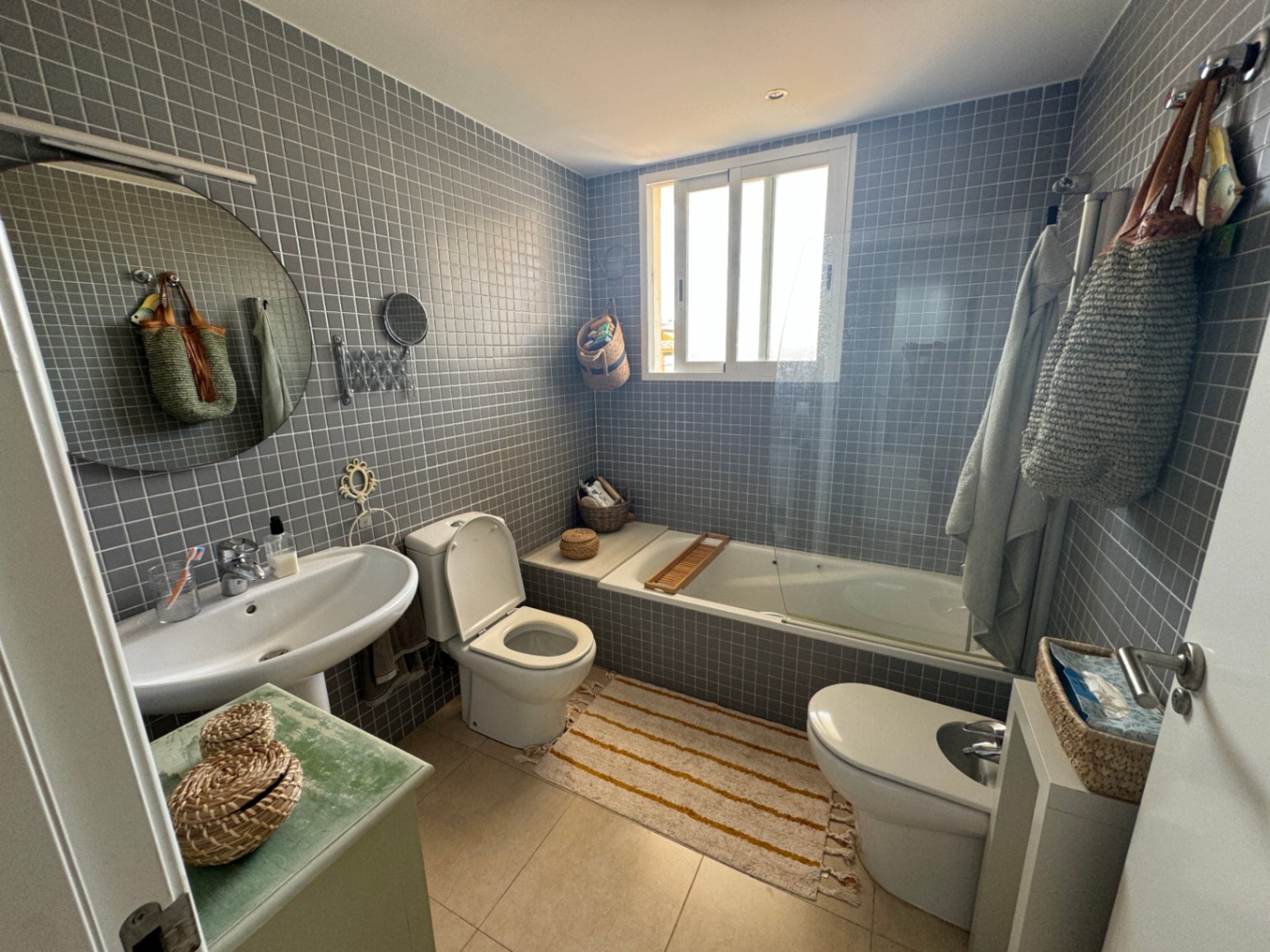 Badkamer met grijze mozaïektegels, voorzien van een witte kolomwastafel, ronde spiegel en een ligbad met glazen douchescherm en natuurlijk daglicht.